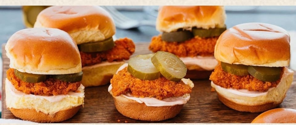 Four crispy chicken sliders with pickles and sauce on brioche buns, on a wooden board.